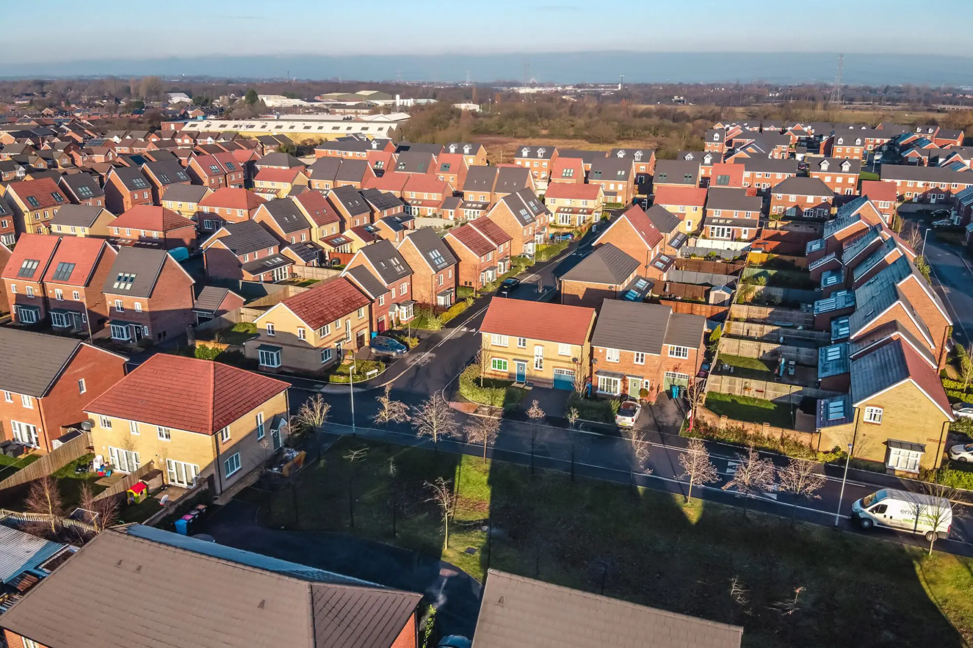 An aerial view of a suburban neighborhood with rows of houses, many of which have solar panels installed on their roofs, surrounded by well-maintained lawns and streets.